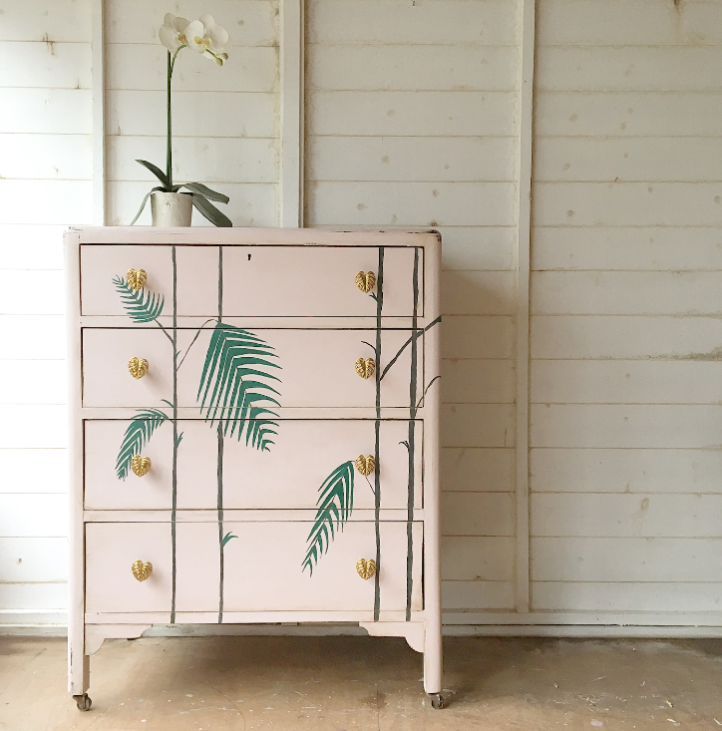 Decorative dresser with palm leaf designs and gold knobs against a wooden wall.