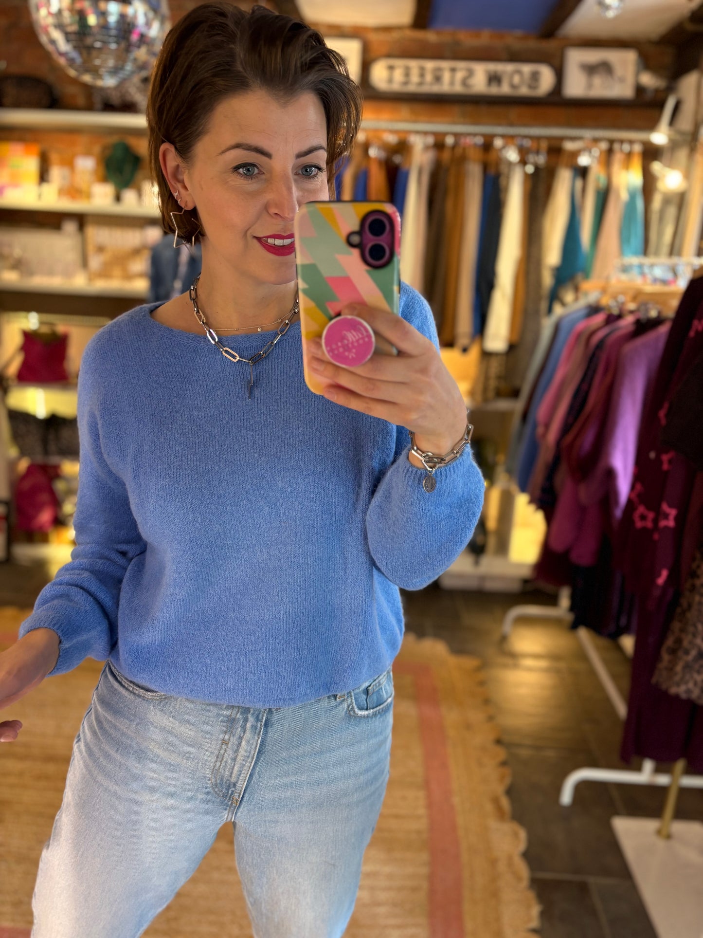 Woman taking a mirror selfie in a clothing store wearing a blue knit jumper and jeans.
