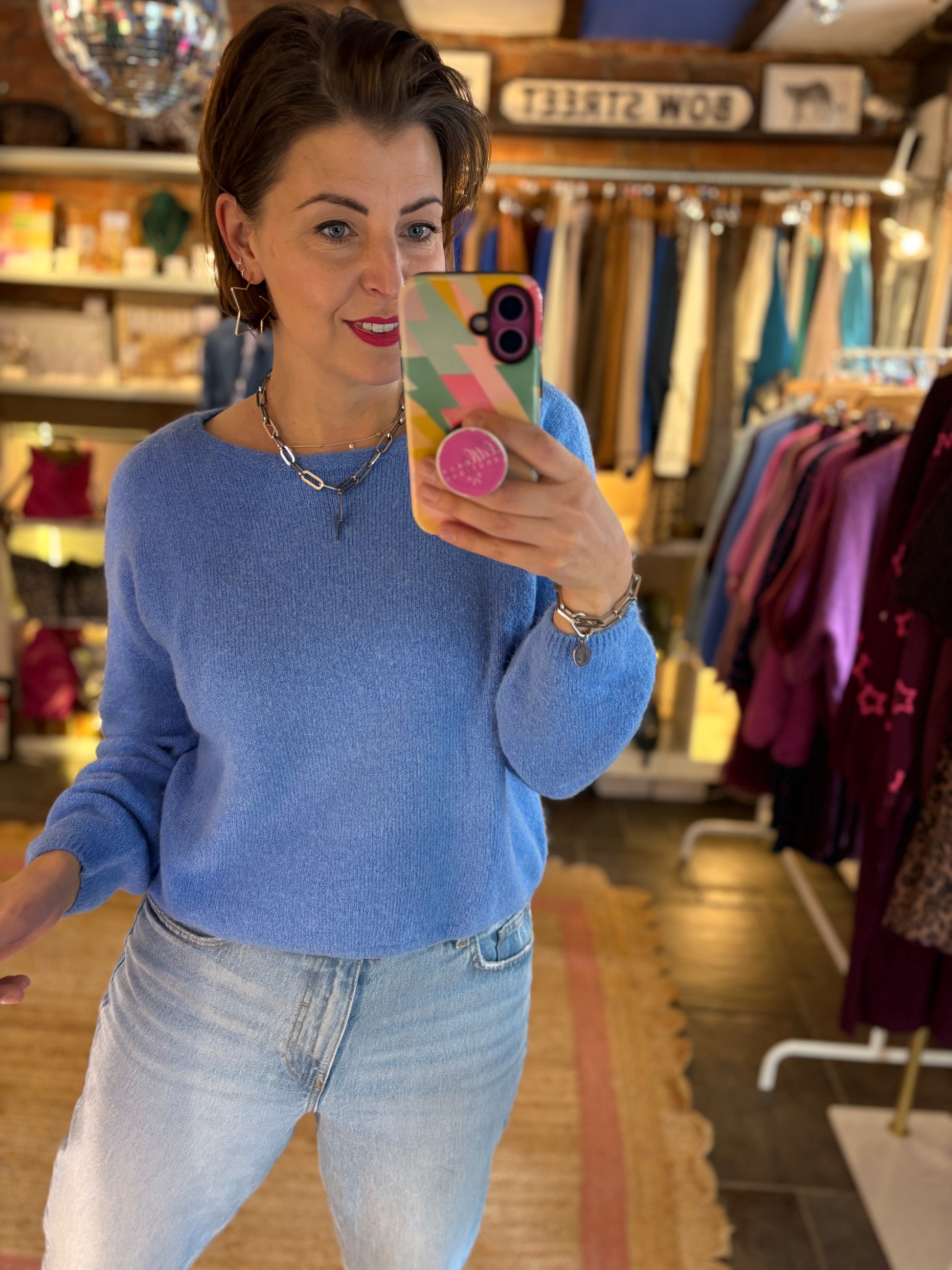 Woman taking a mirror selfie in a clothing store wearing a blue knit jumper and jeans.
