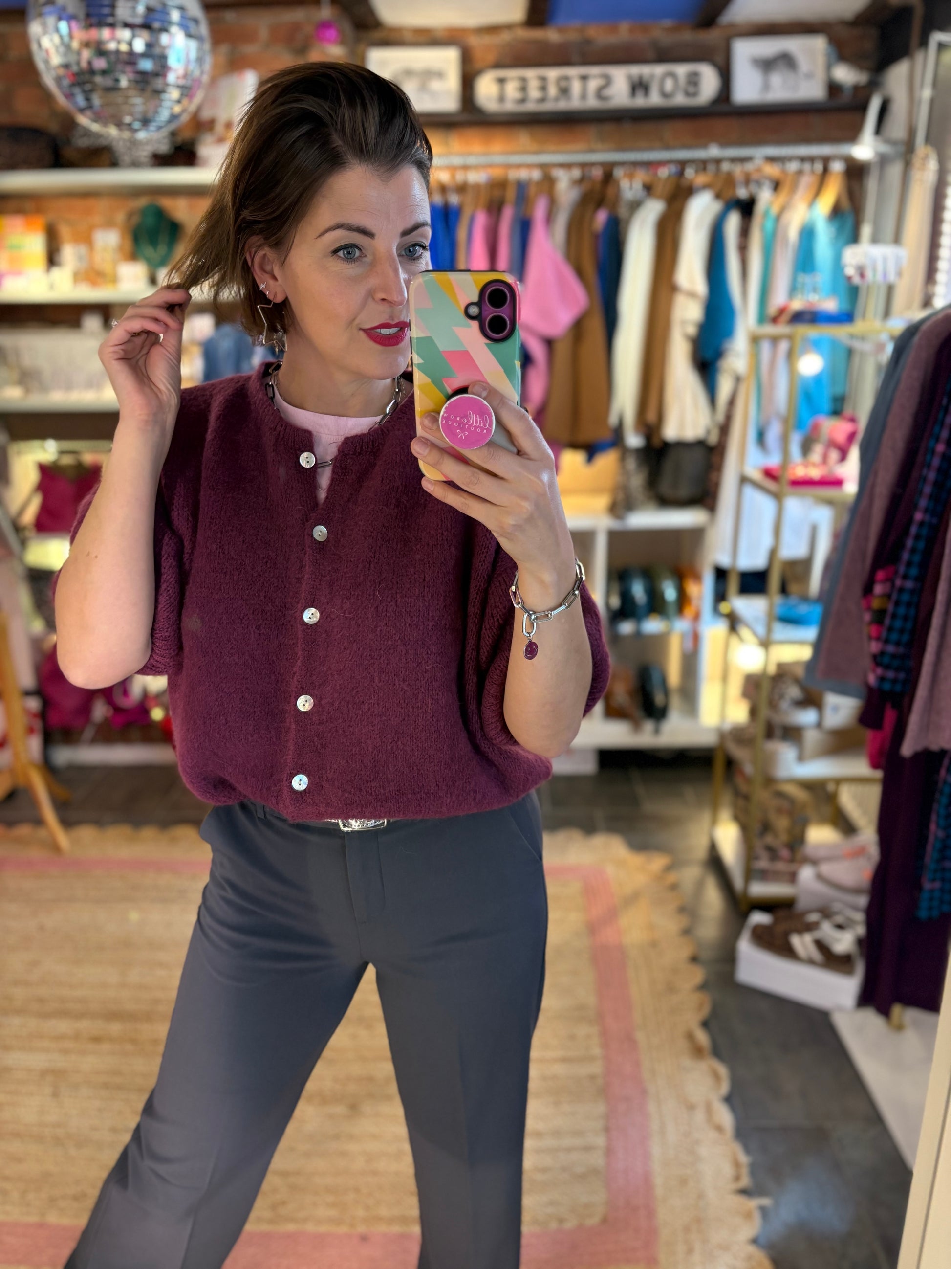 Woman taking a mirror selfie in a clothing store wearing a maroon cardigan and grey trousers