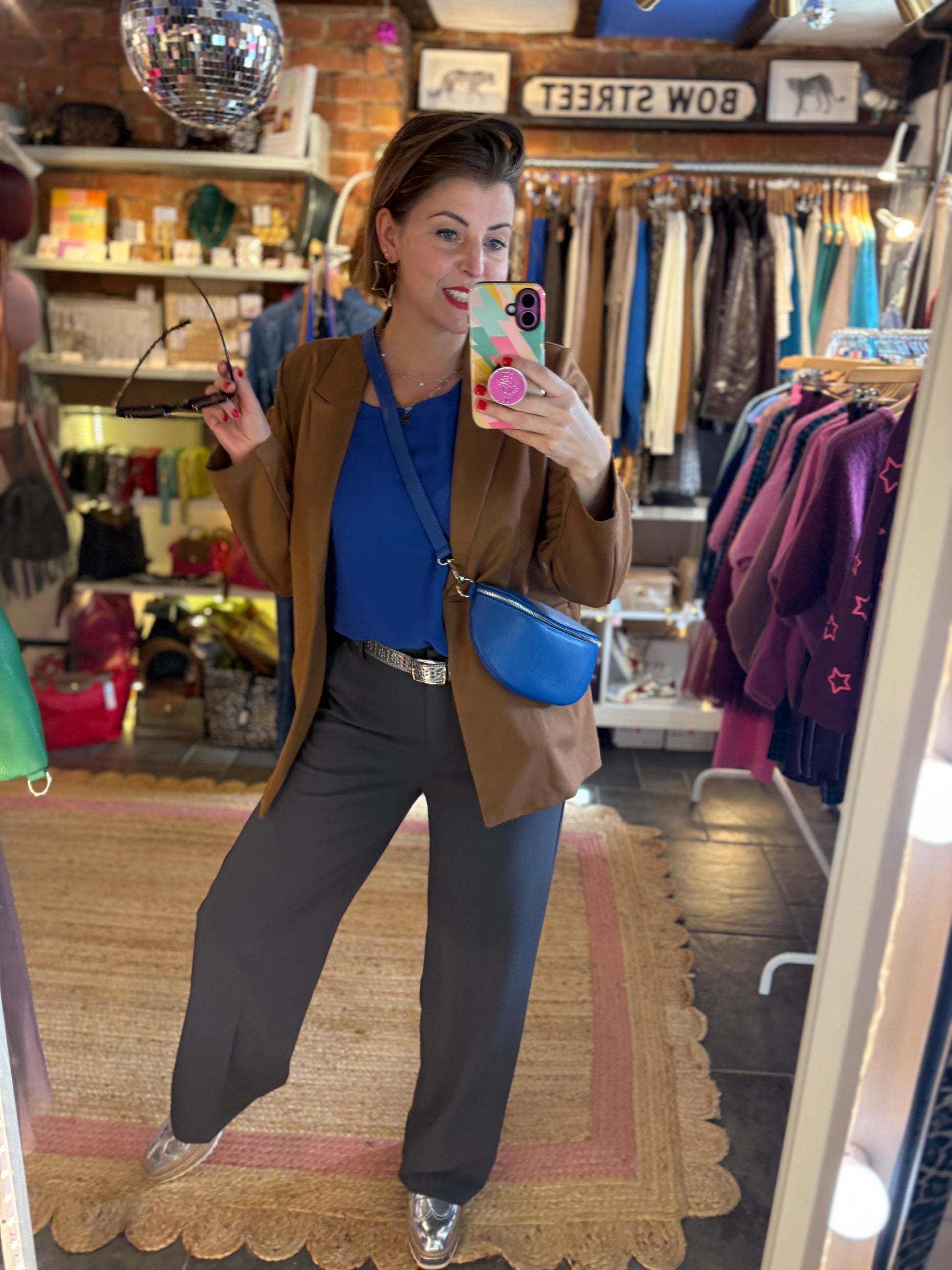 Person taking a mirror selfie in a clothing store wearing a brown jacket, blue top, and grey trousers.