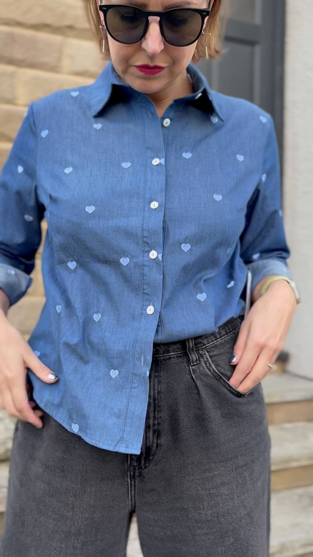 Person wearing a blue denim shirt with heart patterns and dark jeans outdoors.