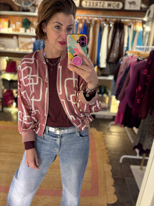 Person taking a mirror selfie wearing a patterned satin bomber jacket and jeans in a clothing store.