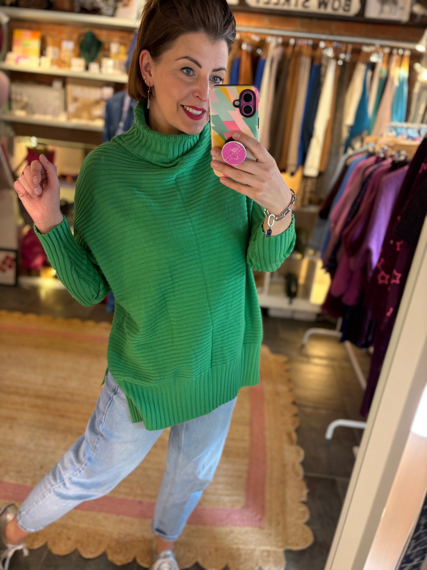 Woman taking a mirror selfie wearing a green ribbed long sweater in a clothing store.