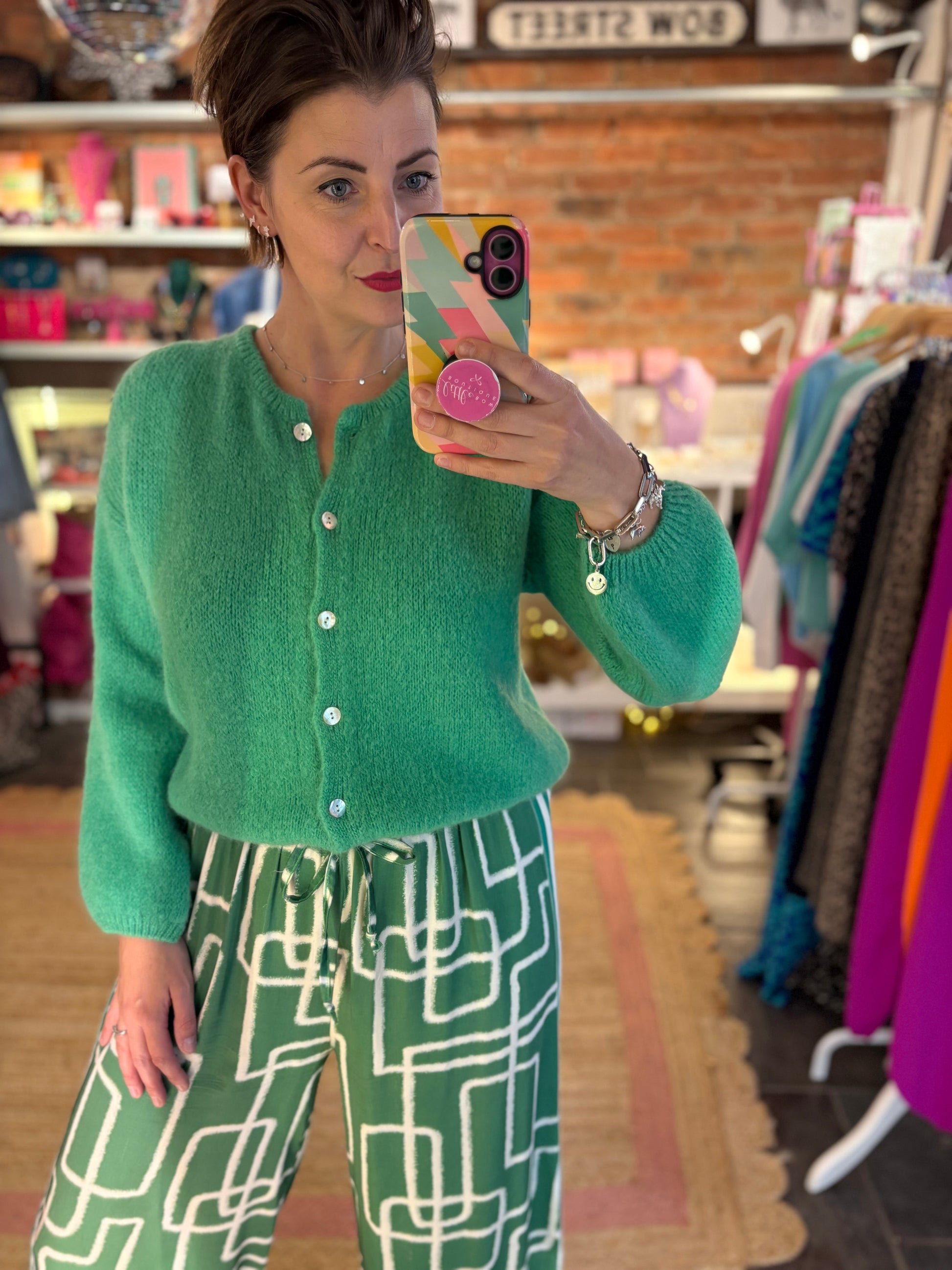 Woman taking a mirror selfie wearing a green cardigan and patterned pants in a store.