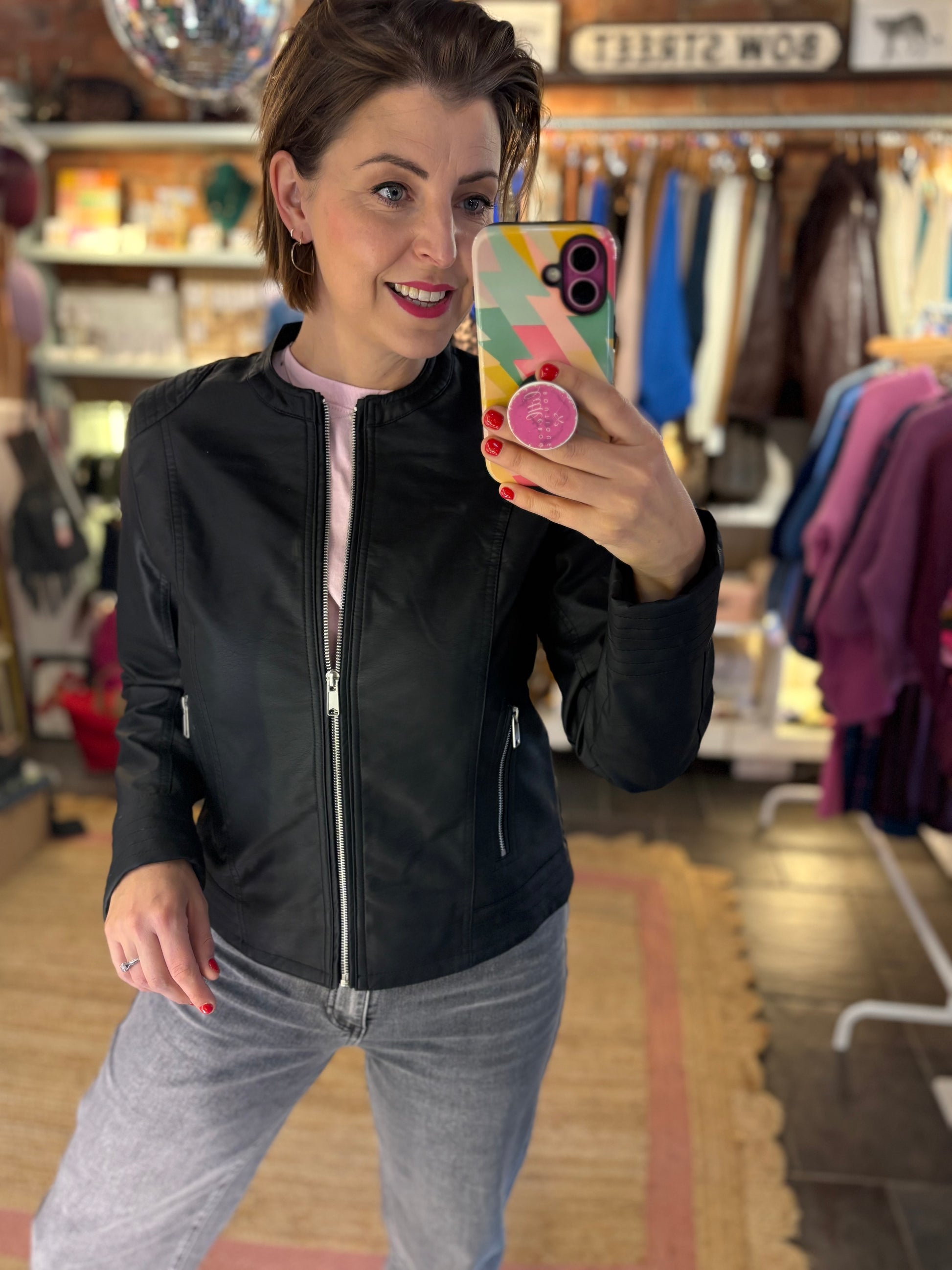 Woman taking a mirror selfie in a clothing store wearing a black collarless jacket and grey pants.