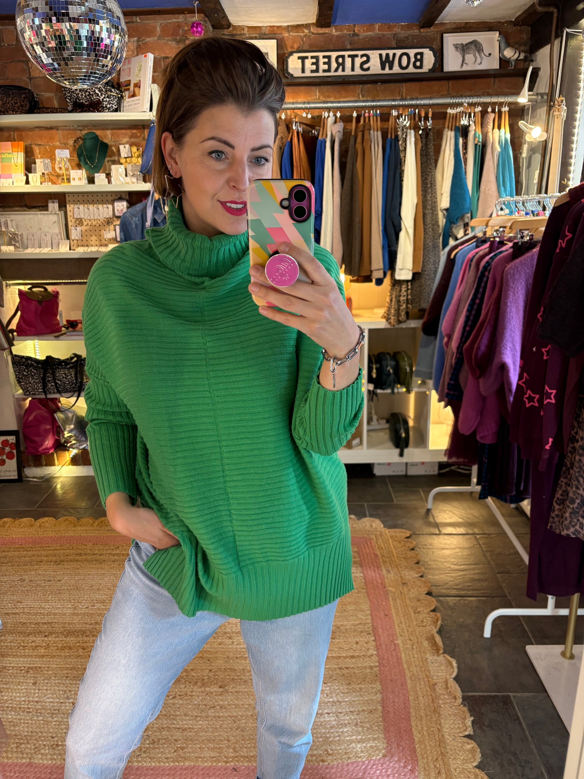 Woman in a green ribbed sweater taking a selfie in a store with light blue jeans