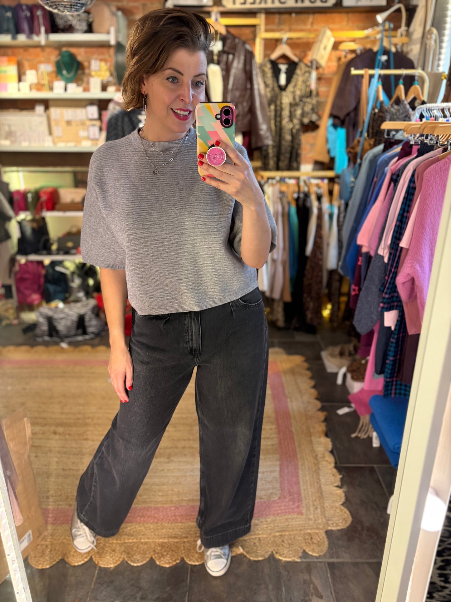 Person taking a mirror selfie wearing a gray sweater and black pants in a clothing store.