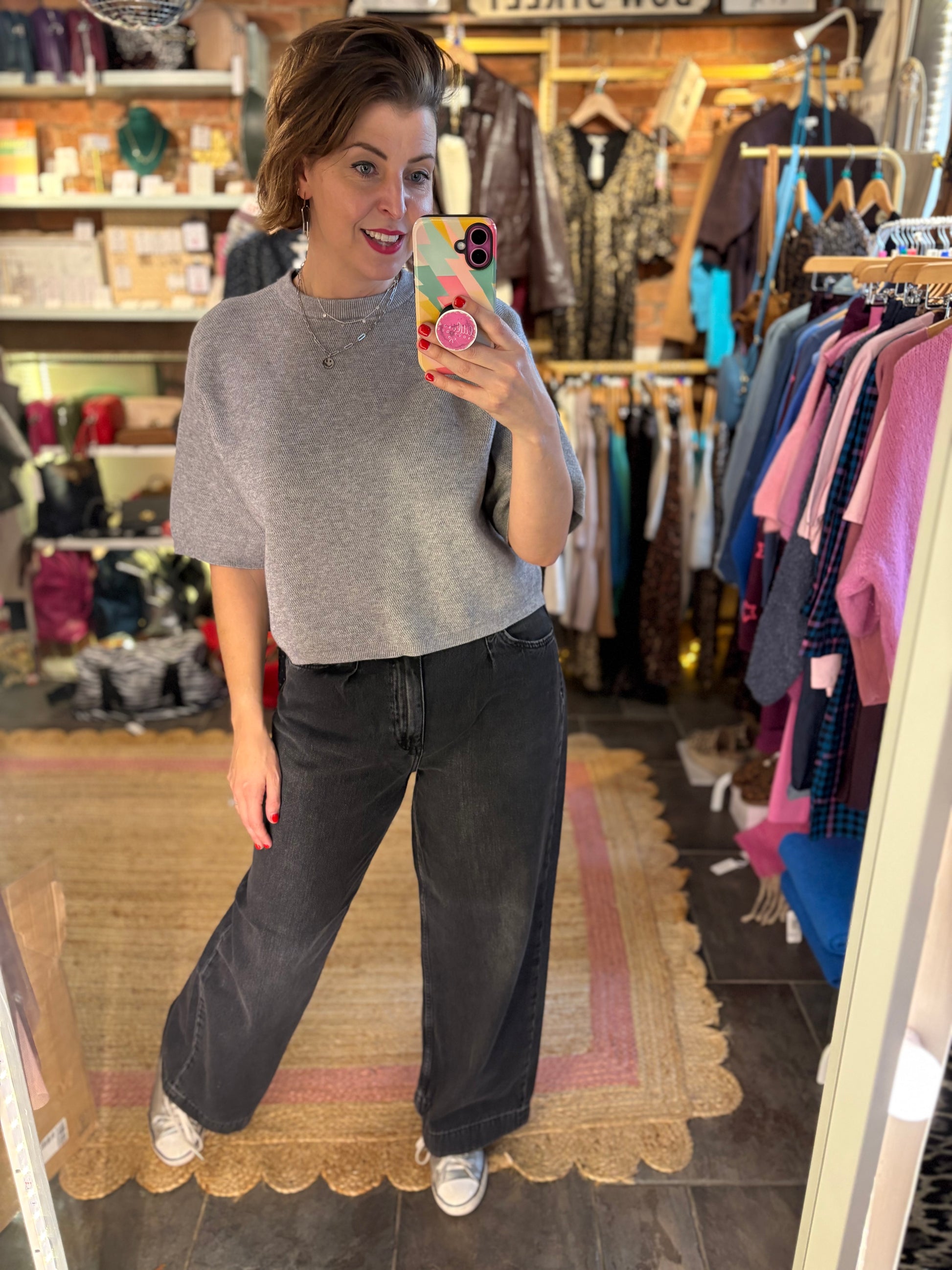 Person taking a mirror selfie wearing a gray sweater and black pants in a clothing store.
