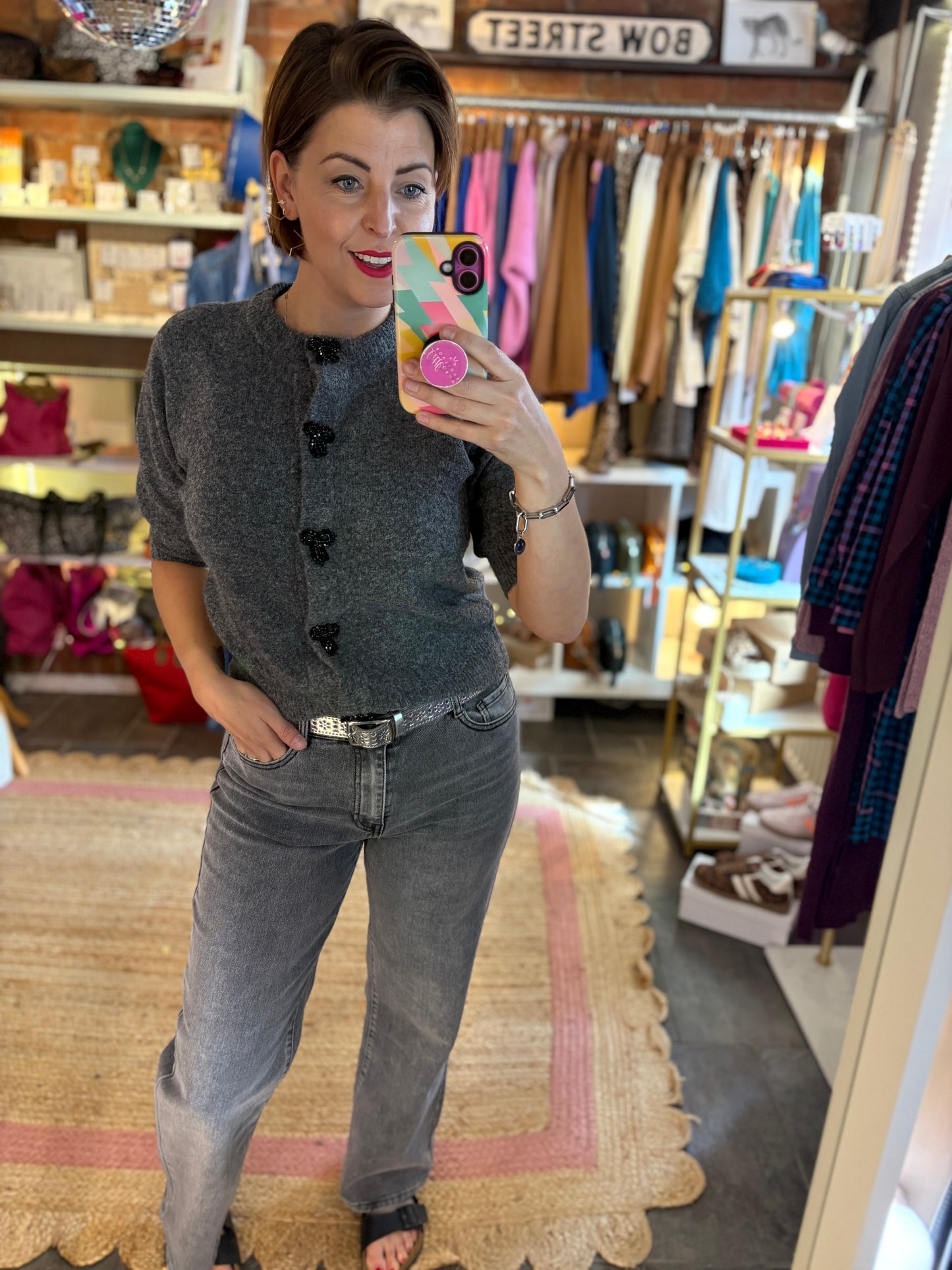 Woman taking a mirror selfie in a clothing store wearing a grey knitted cardigan top and jeans.