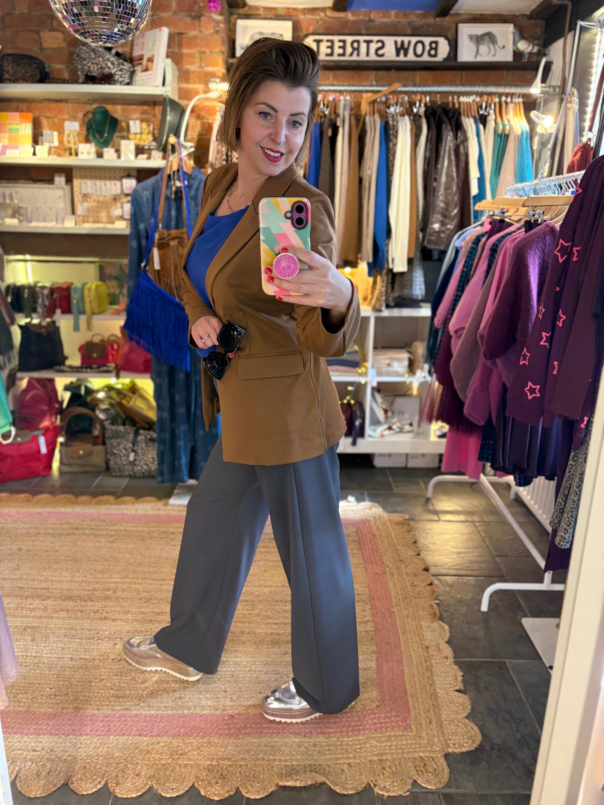 Woman taking a mirror selfie in a clothing store wearing a toffee brown blazer and grey pants.
