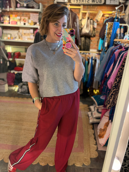 Woman taking a mirror selfie in a clothing store wearing a gray sweater and red pants.