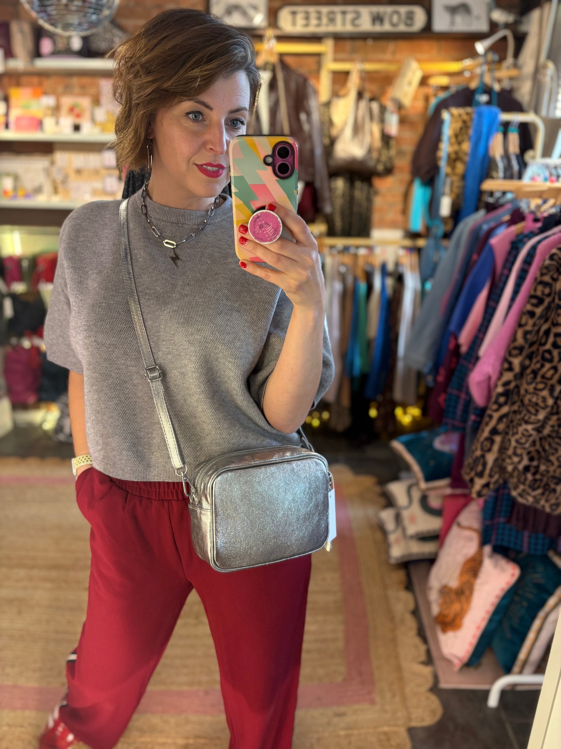 Woman taking a mirror selfie in a clothing store wearing a gray sweater and red pants with a silver crossbody bag.