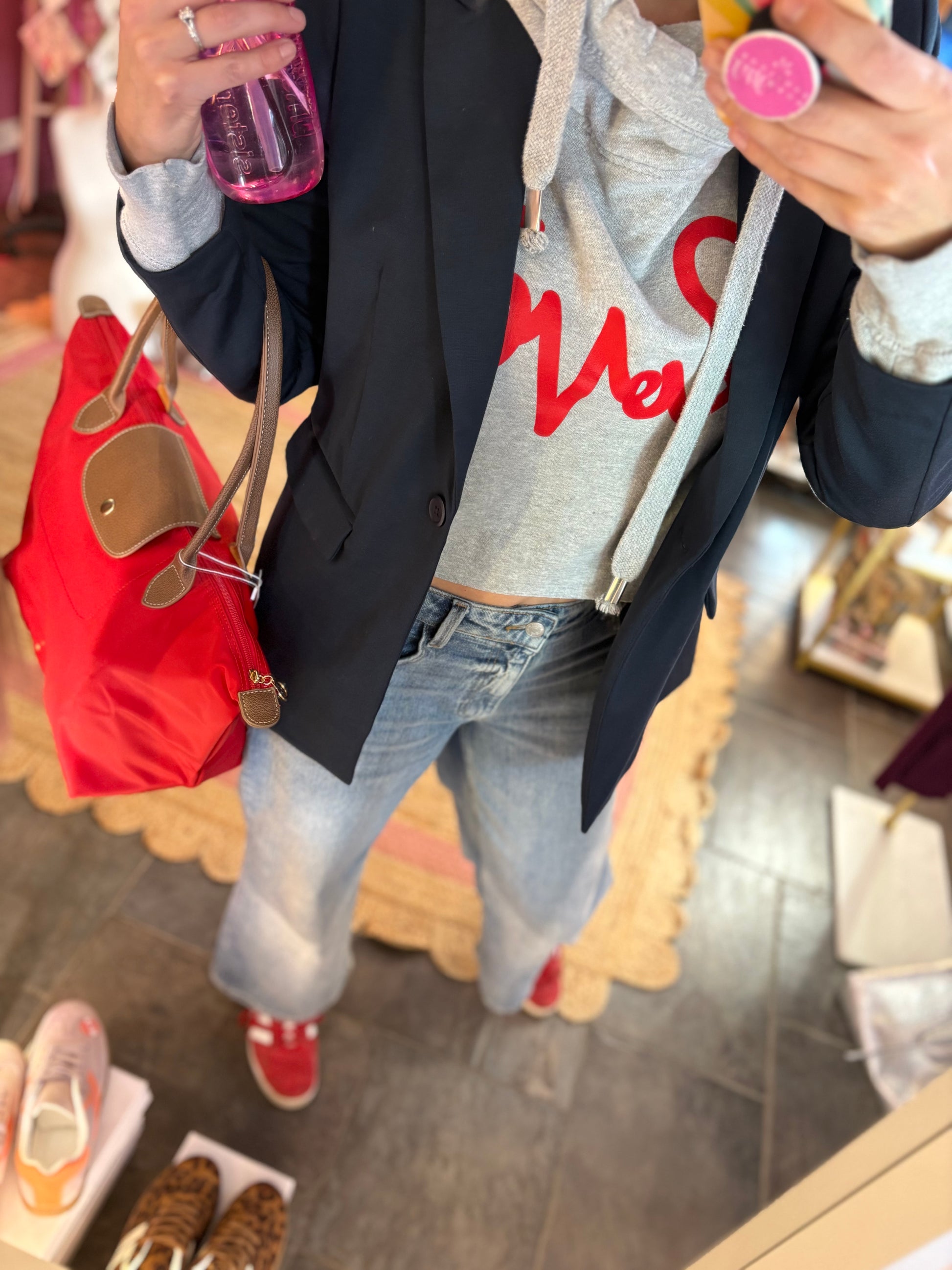 Person wearing a gray hoodie with red text, a red bag and navy blue jacket, and jeans in a store setting.