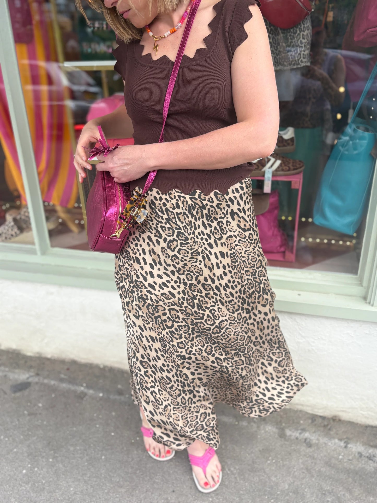 Woman wearing a leopard print skirt and brown top, holding a pink bag, standing on a street.