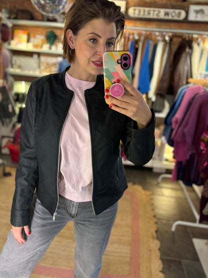 Woman taking a mirror selfie in a clothing store wearing a black collarless jacket and pink tshirt.