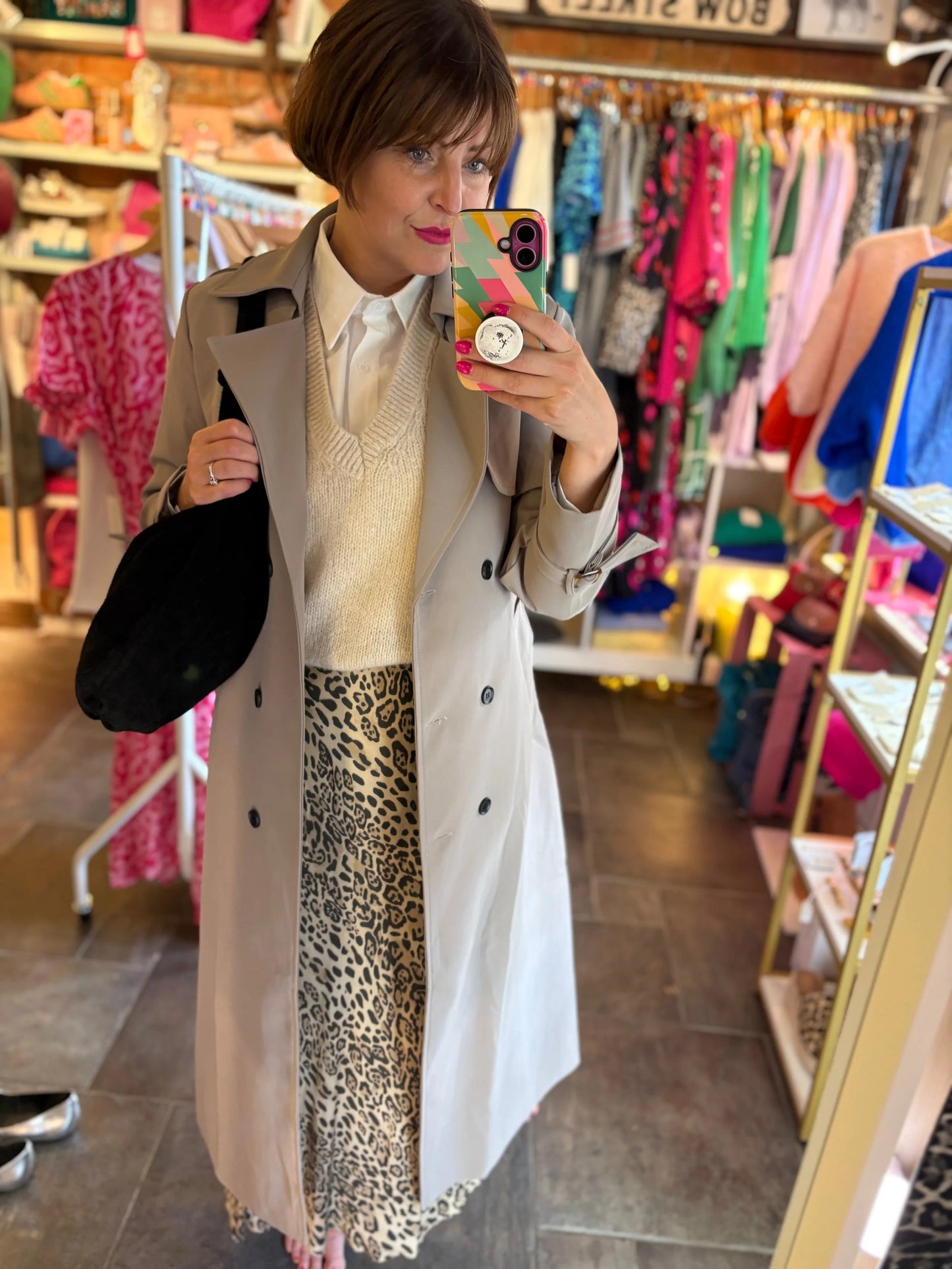 Person taking a mirror selfie in a clothing store wearing a trench coat and leopard print skirt.