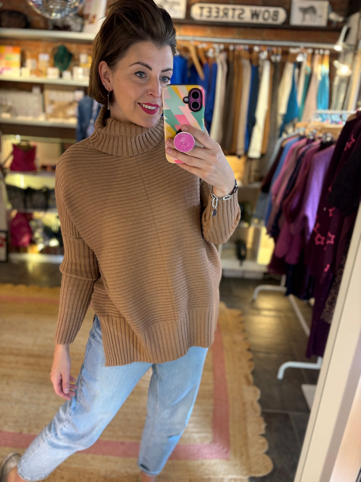 Woman taking a mirror selfie wearing a camel brown sweater and light blue jeans in a clothing store.
