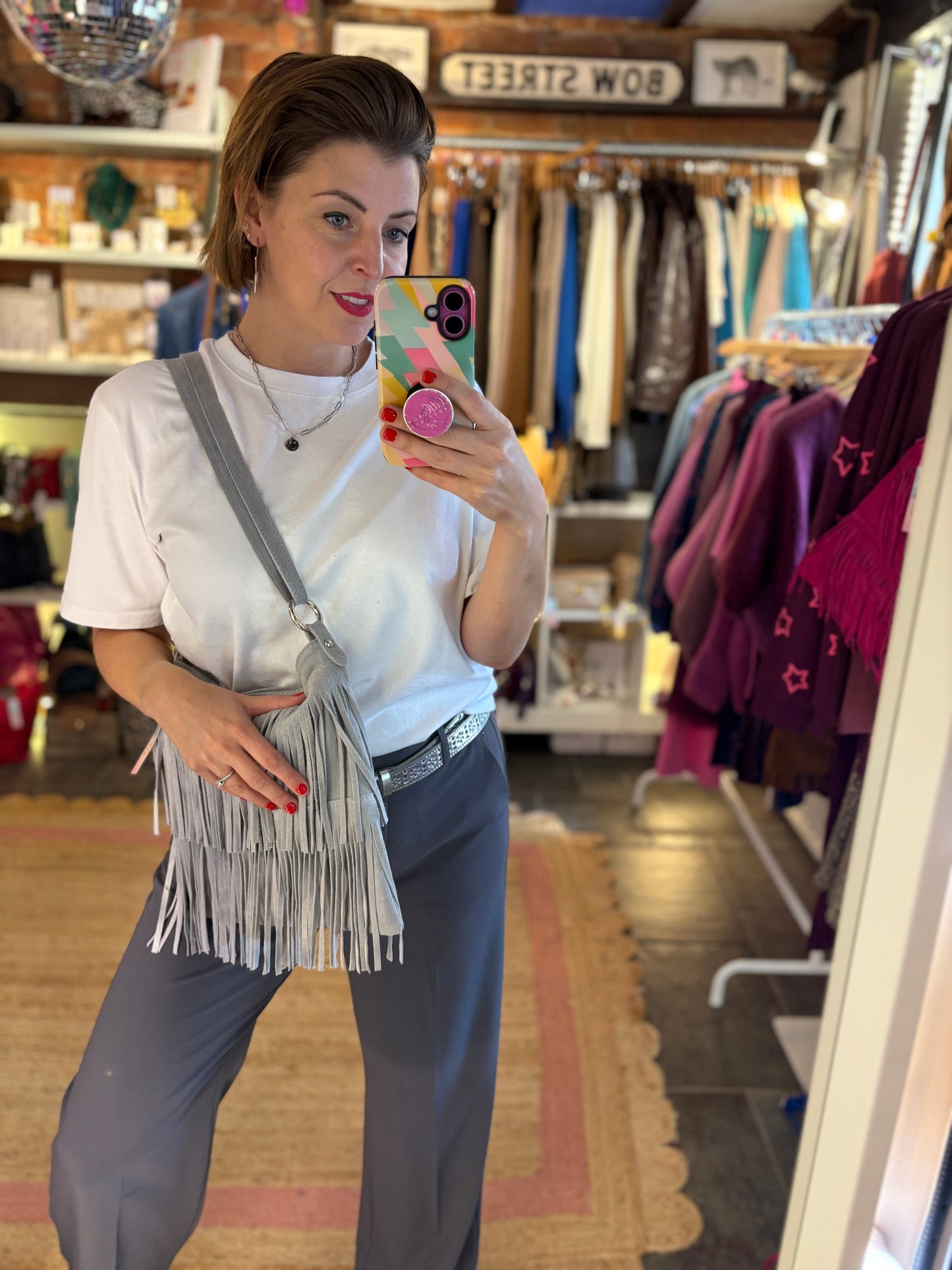 Woman taking a mirror selfie in a clothing store wearing a white shirt, grey trousers, and a fringed suede grey bag.