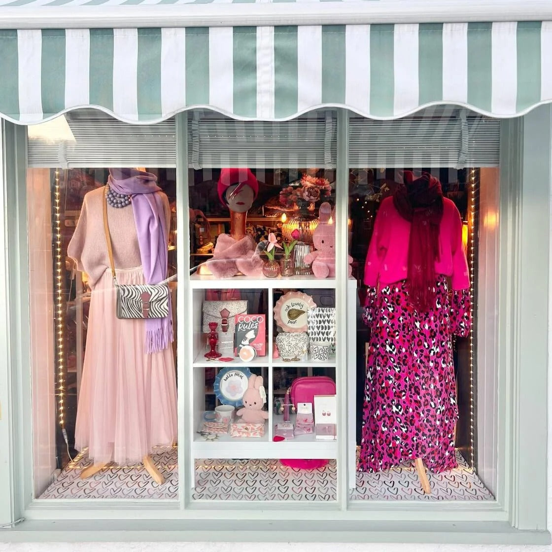 Store window display with mannequins wearing pink and leopard print dresses under a striped awning.