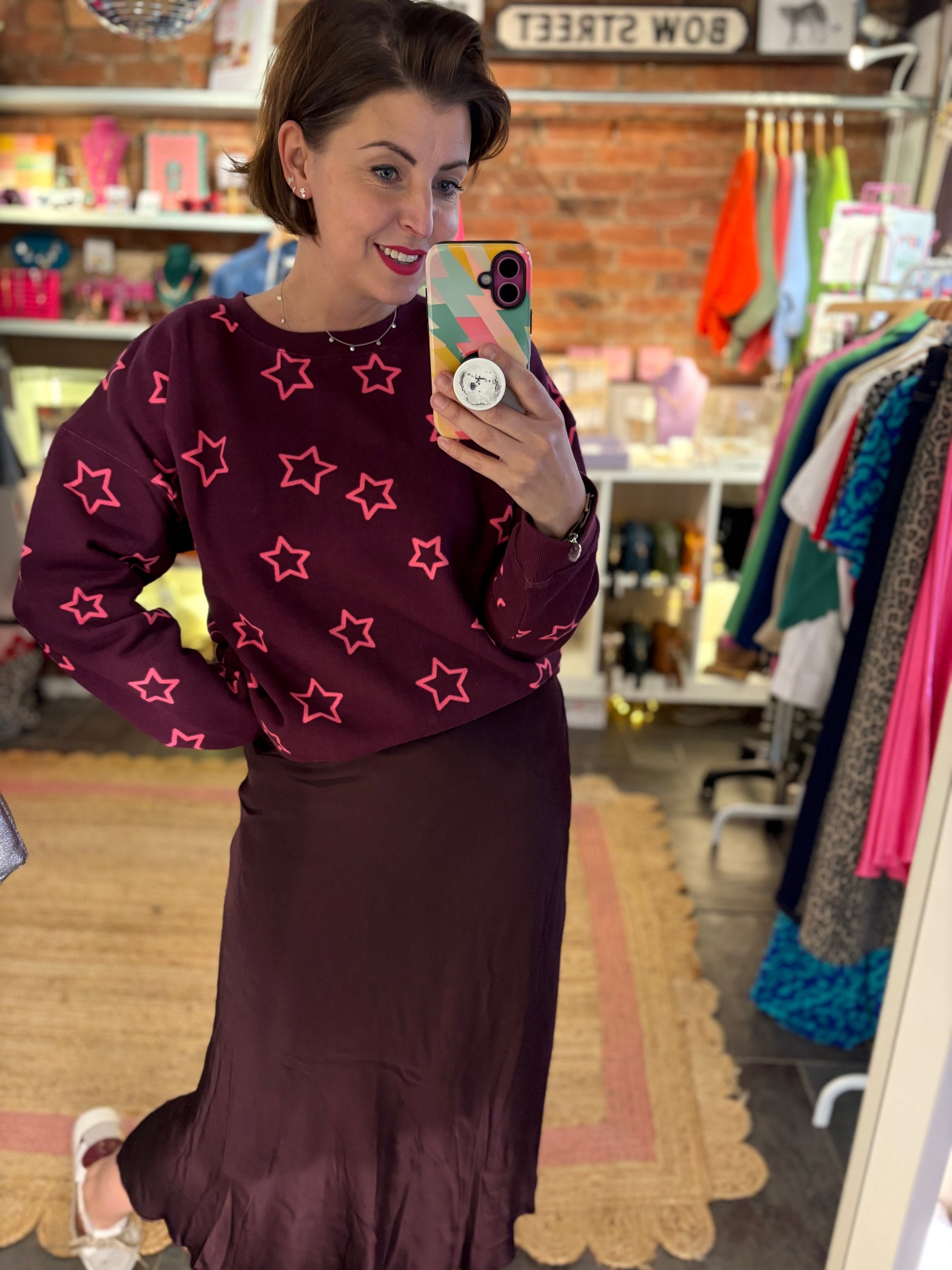 Woman taking a mirror selfie wearing a maroon sweater with pink star patterns and a matching skirt in a store.