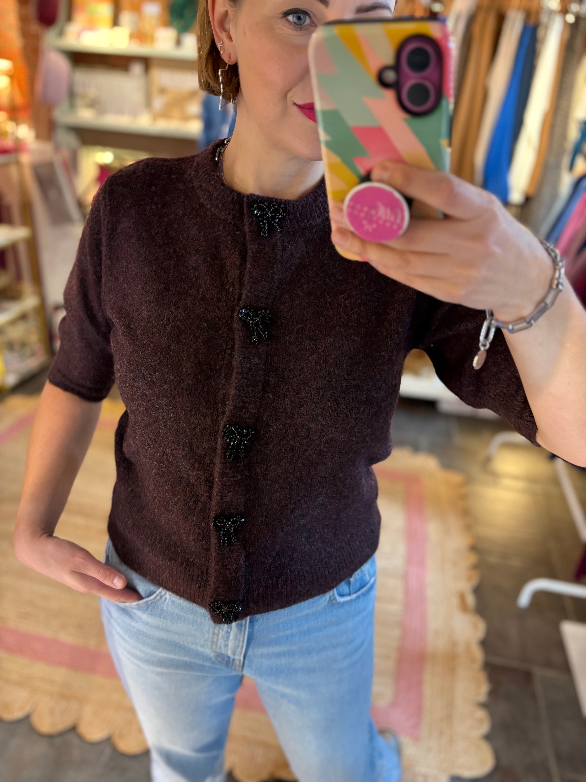 Person taking a mirror selfie wearing a merlot burgundy cardigan and light blue jeans in a store setting.
