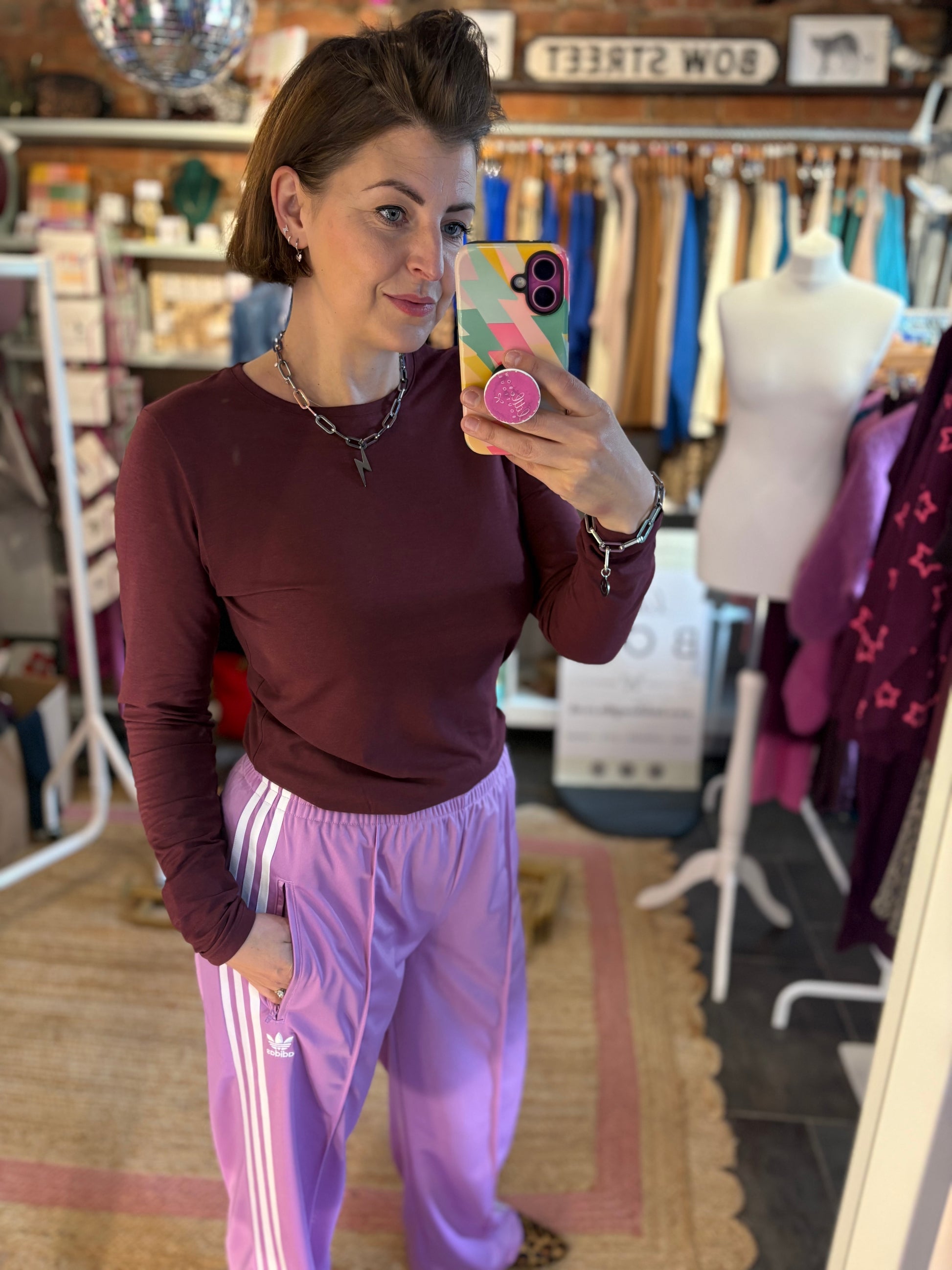 Woman taking a mirror selfie in a clothing store wearing a maroon top and purple tracksuit bottoms