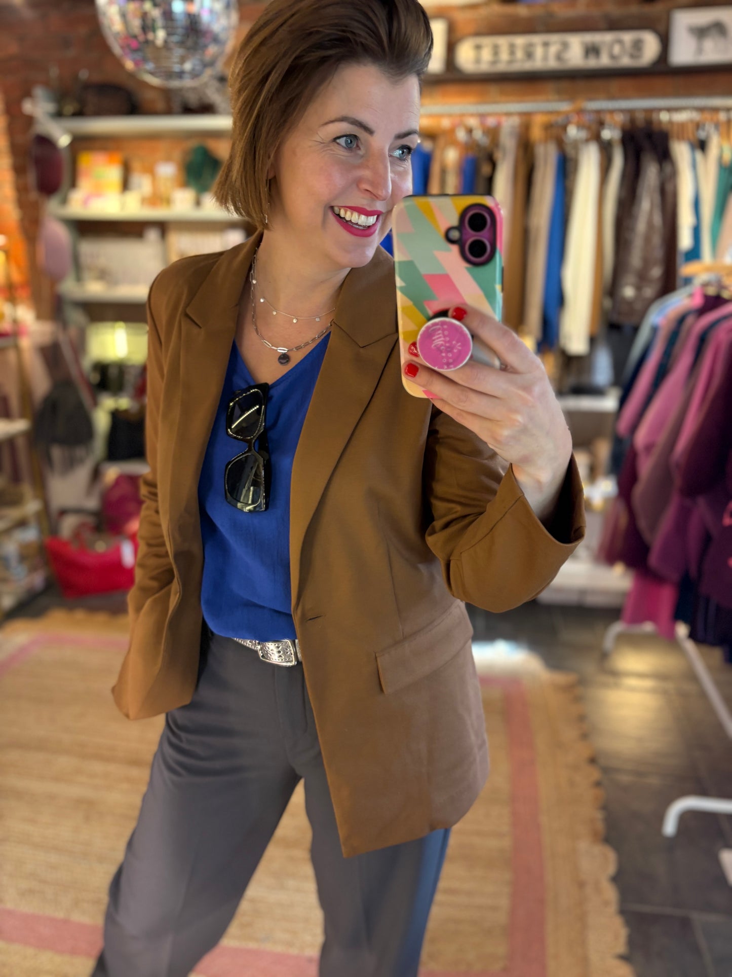 Woman taking a mirror selfie in a clothing store wearing a brown blazer and blue shirt.