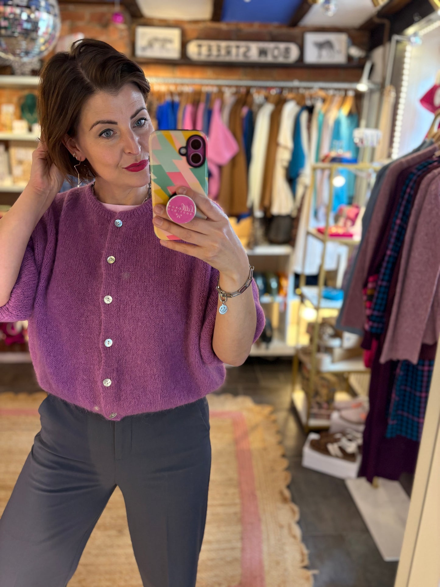 Woman taking a mirror selfie wearing a purple cardigan in a clothing store.