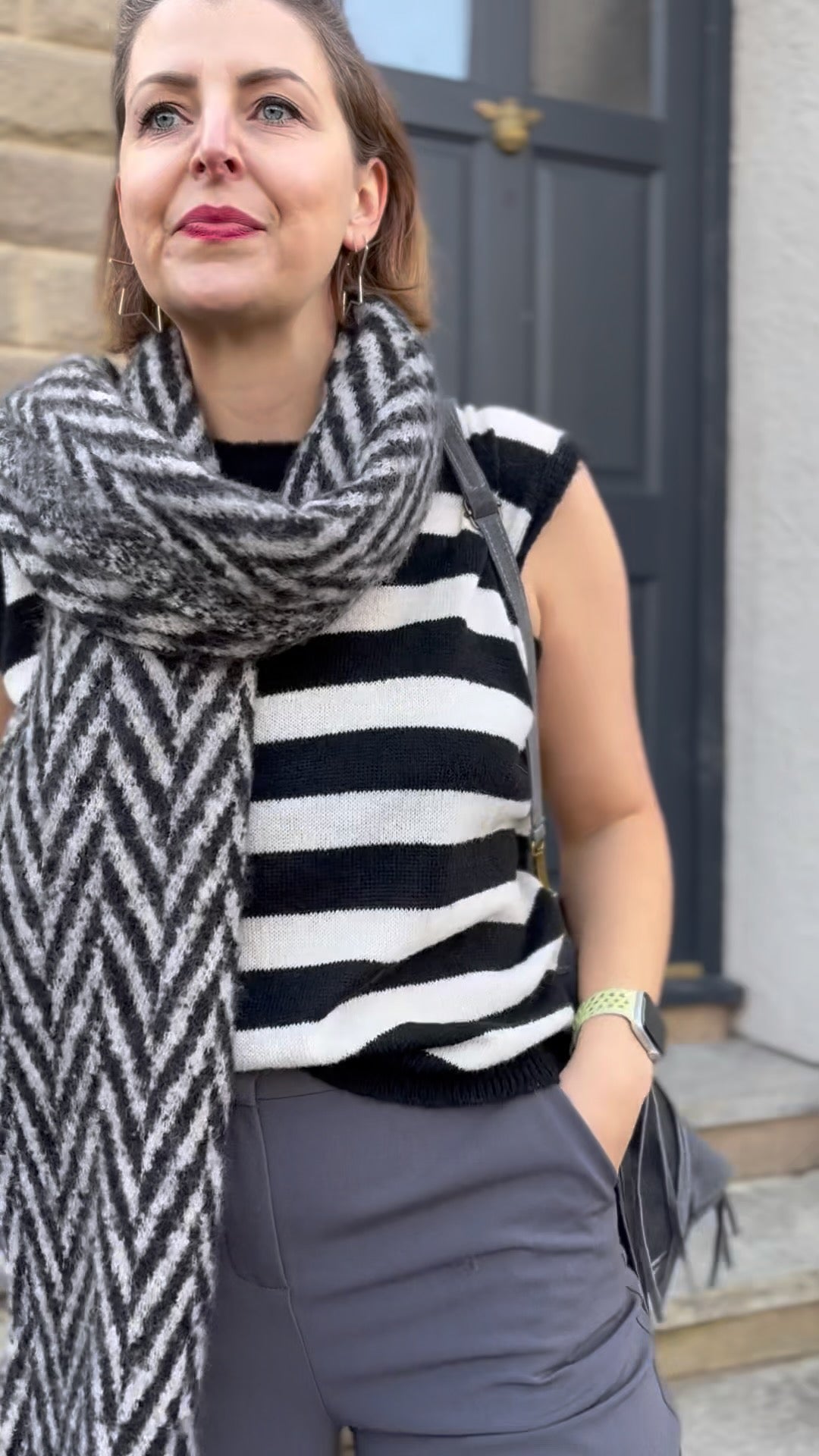 Woman wearing a black and white striped sleeveless top and a large scarf, standing in front of a door.