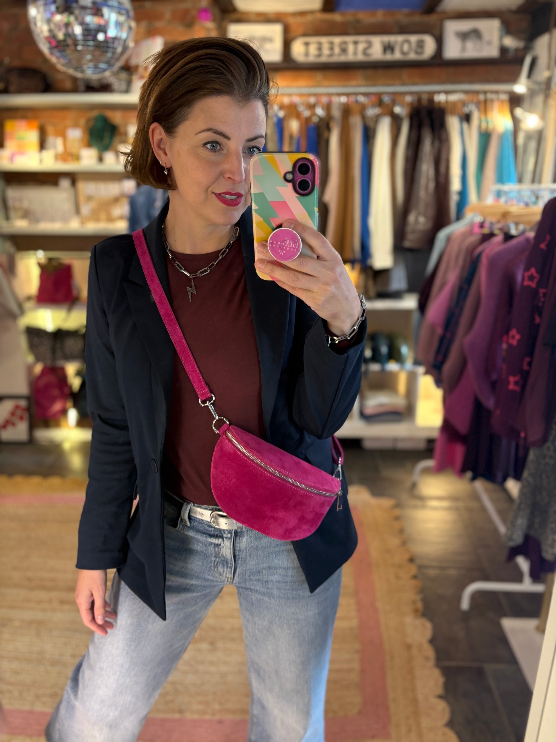 Woman taking a mirror selfie in a clothing store wearing a navy blazer jacket, blue denim jeans and a suede pink crossbody bag.
