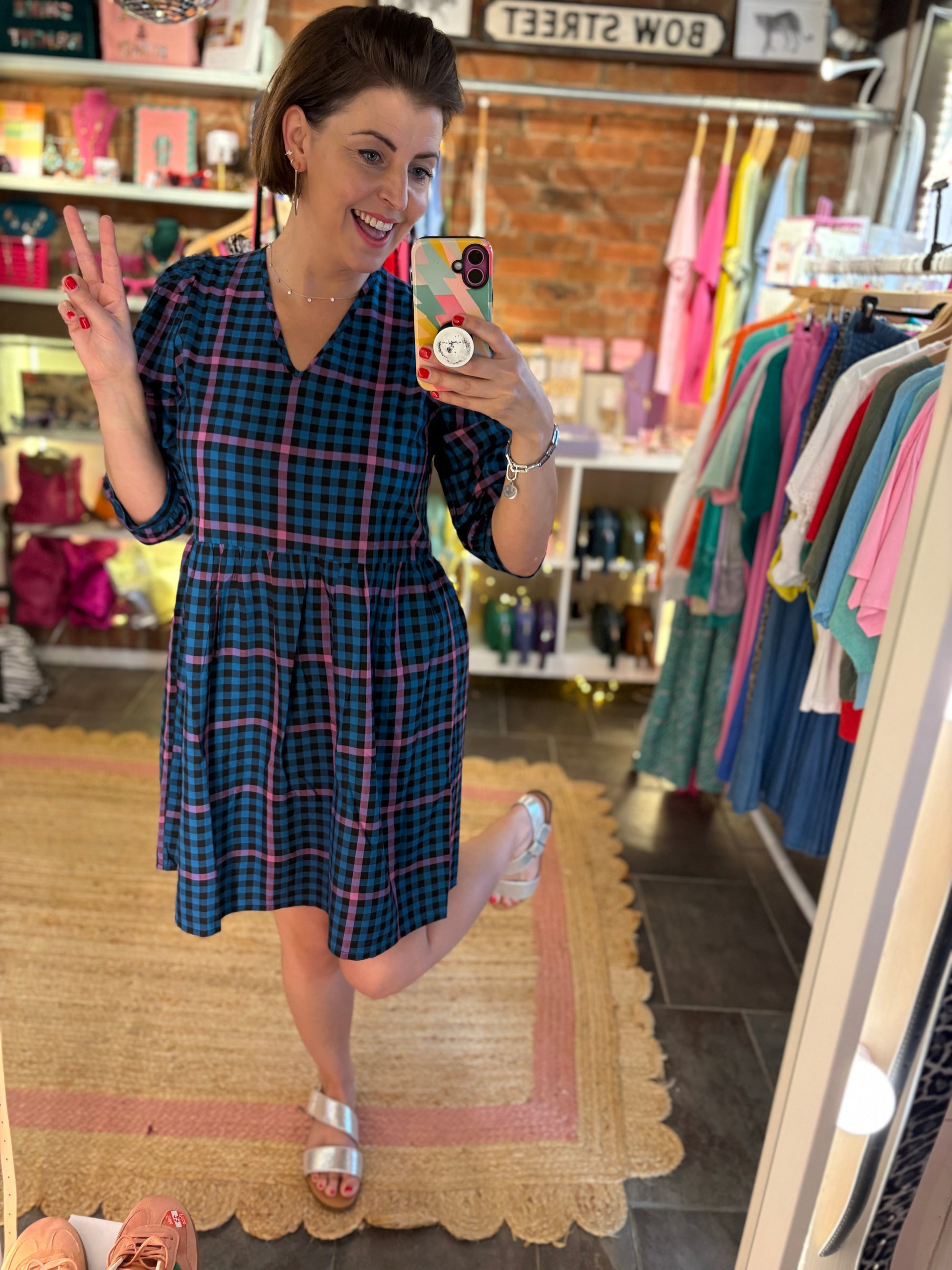 Woman taking a mirror selfie wearing a blue plaid dress in a store.