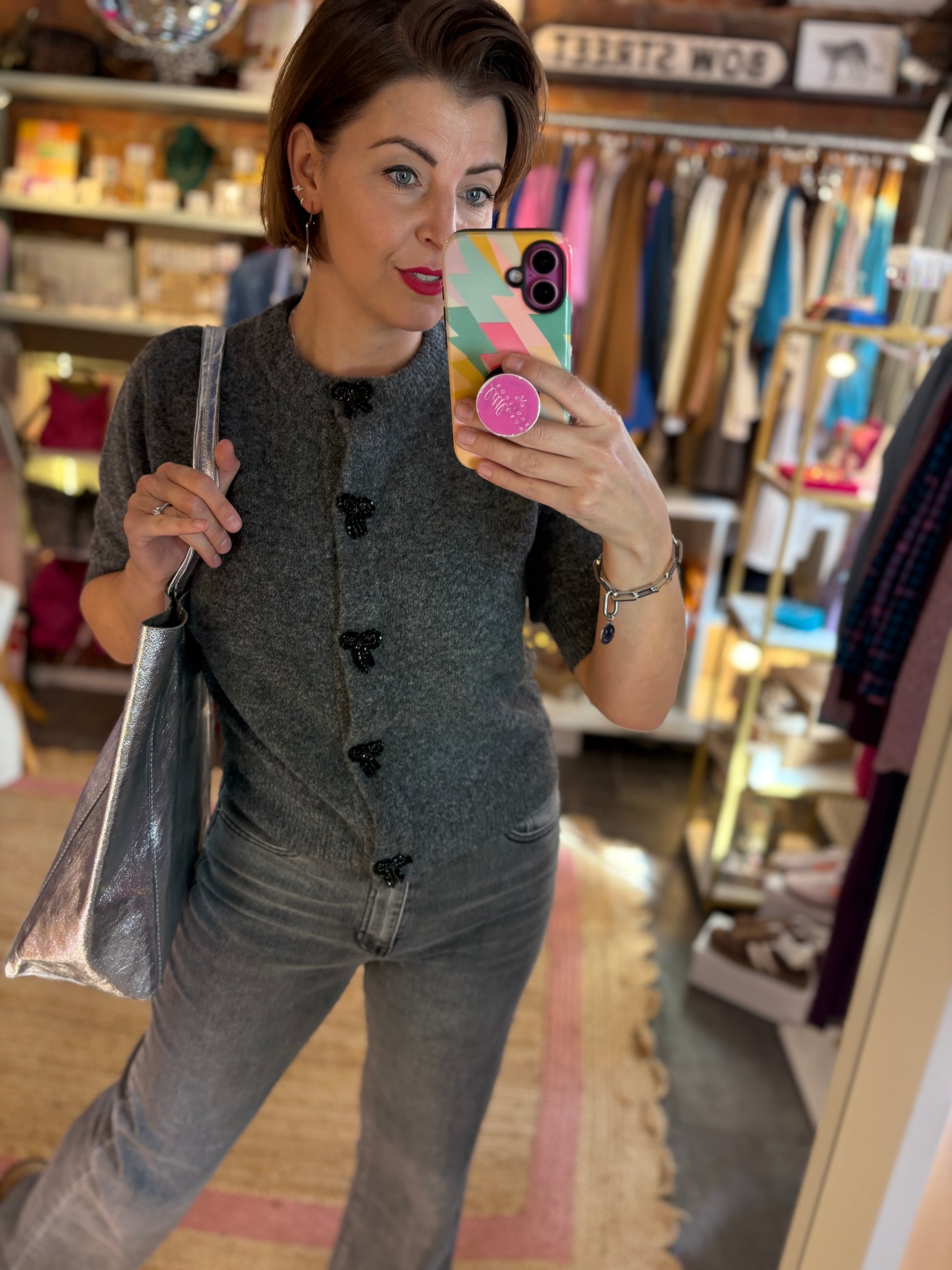 Woman taking a mirror selfie in a clothing store wearing a grey outfit with black sparkle bow button detailing