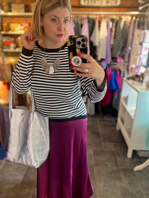 Woman taking a mirror selfie in a store wearing a striped sweater and purple skirt holding a silver metallic tote bag.
