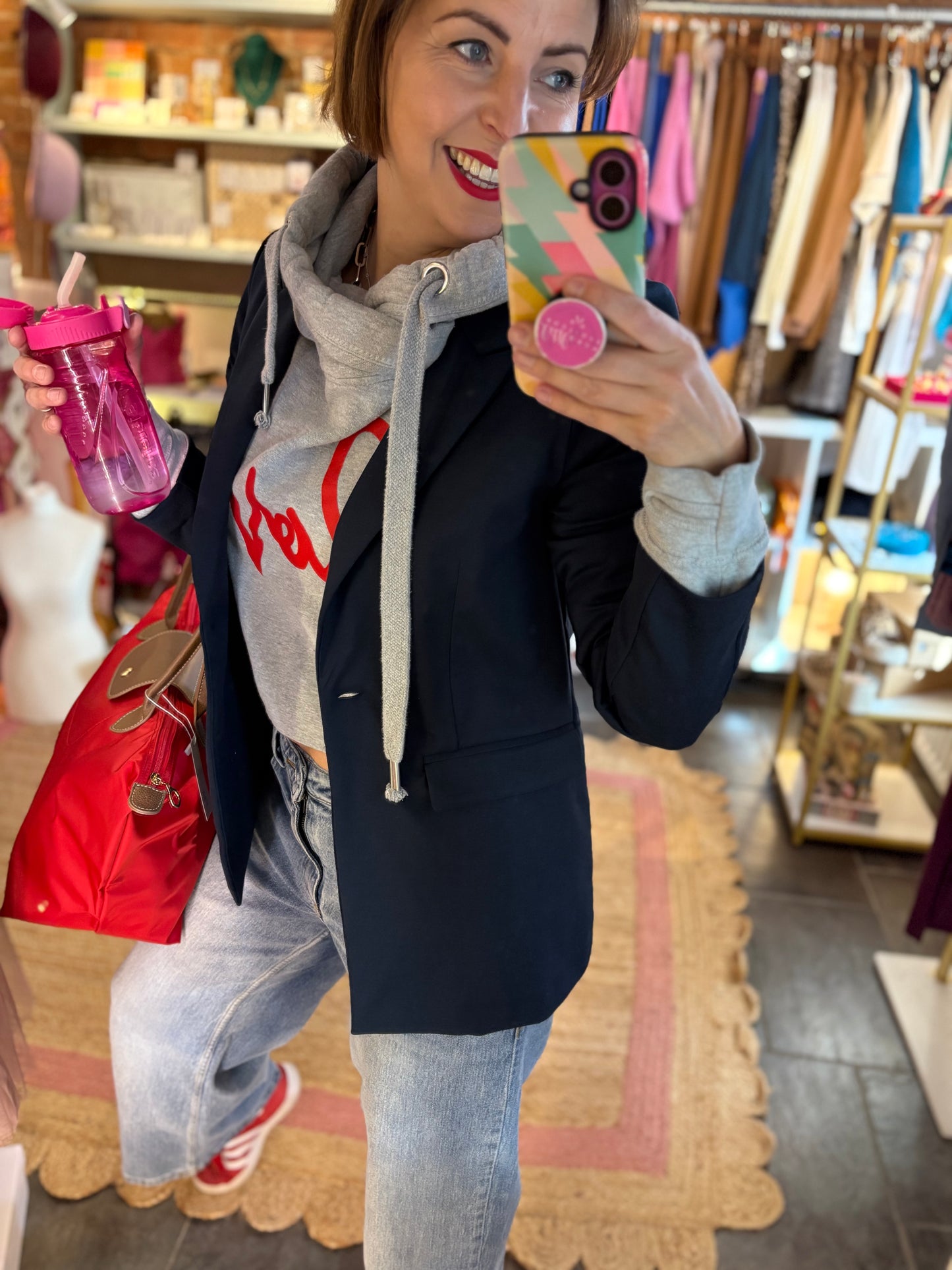 Woman taking a mirror selfie in a store, wearing a gray hoodie, blue jeans and a red bag. A smart casual look