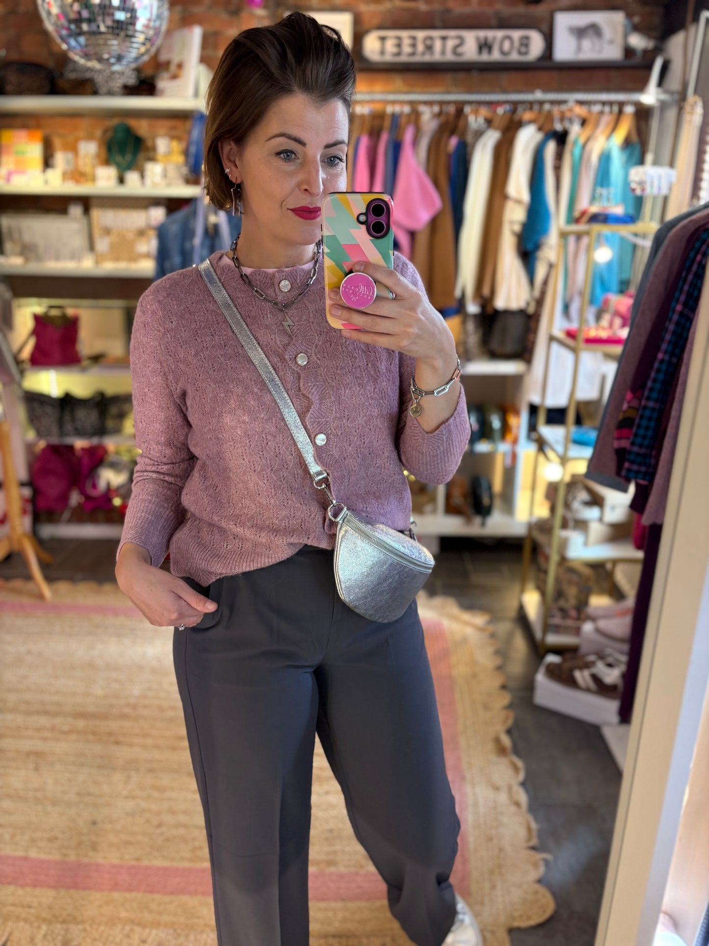 Woman taking a mirror selfie in a clothing store wearing a soft pink cardigan and grey pants.