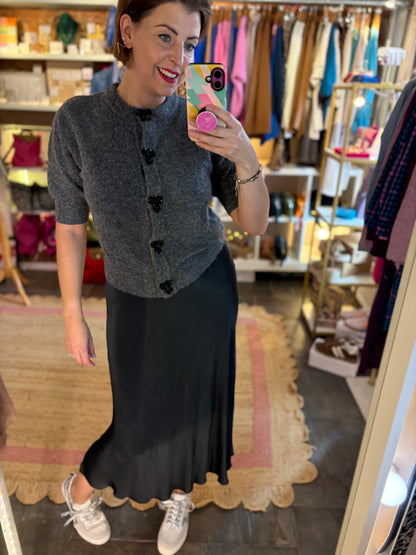 Woman taking a mirror selfie in a clothing store wearing a gray cardigan and black satin slip skirt.