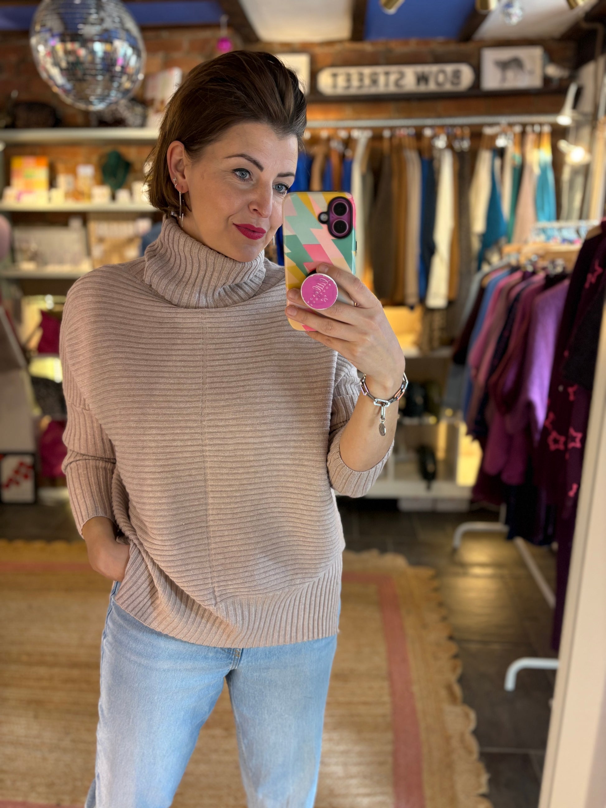 Woman taking a mirror selfie wearing a beige turtleneck sweater and light blue jeans in a clothing store.