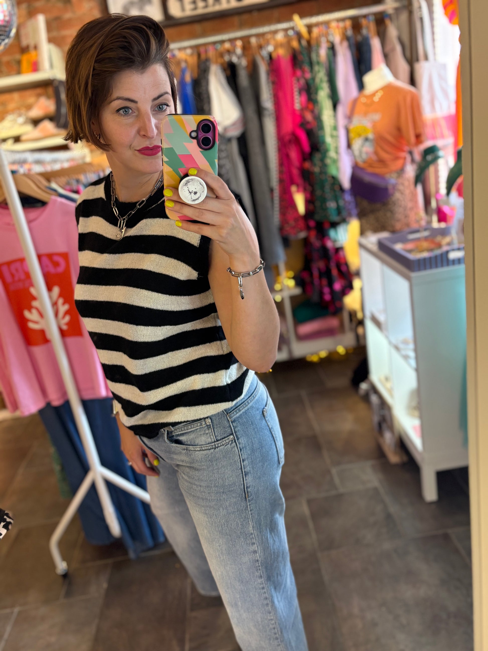 Person taking a mirror selfie wearing a striped knitted sleeveless jumper and jeans in a clothing store.