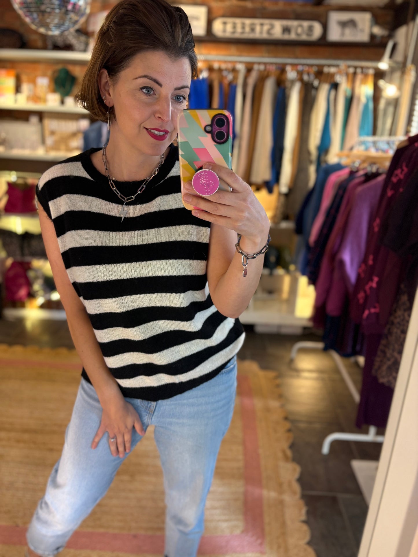 Woman taking a mirror selfie wearing a black and white striped sleeveless top and blue jeans in a clothing store.