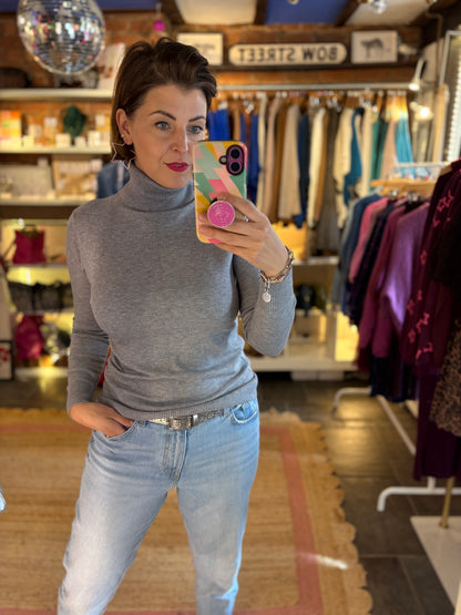 Woman taking a mirror selfie in a clothing store wearing a grey roll neck jumper and light blue jeans.